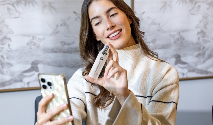 A woman taking a phone selfie with a Neora IllumaBoost bottle.