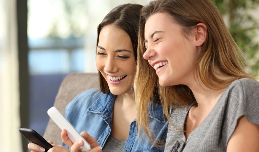 Two women smiling while looking at their phones.