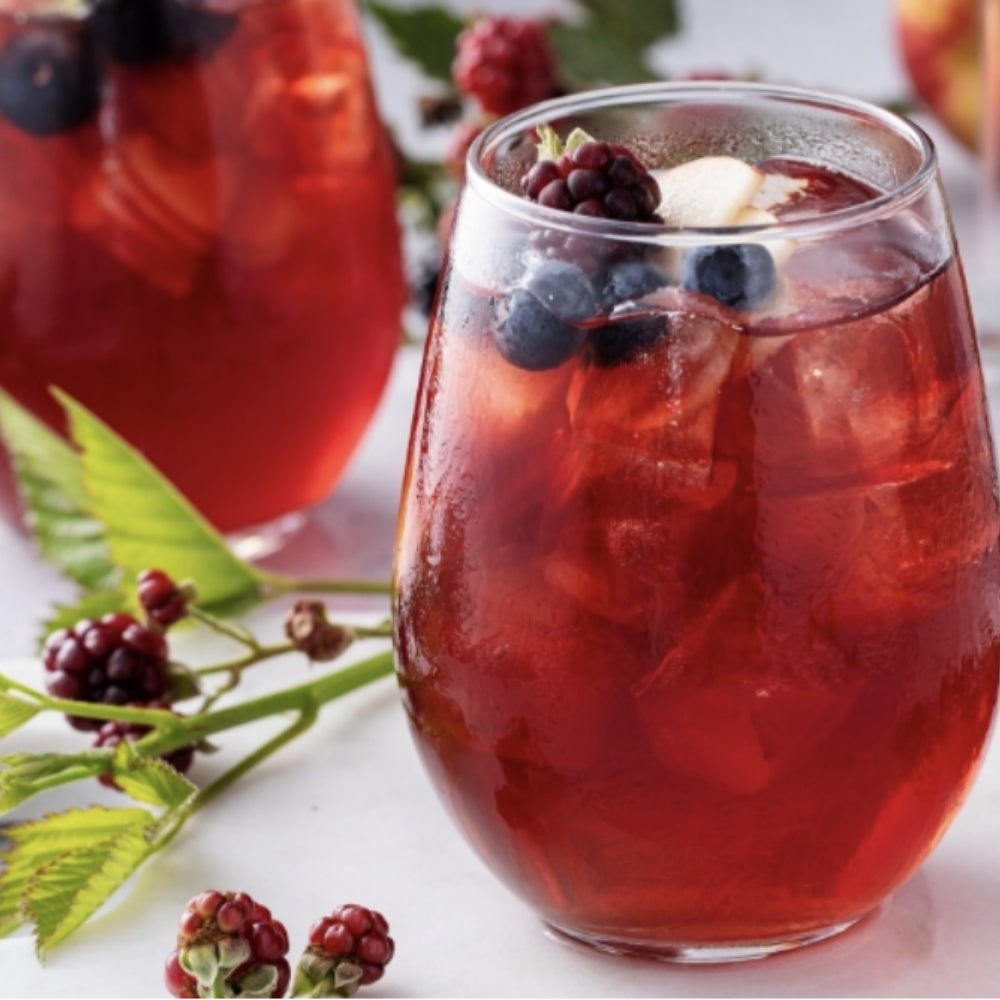 A glass filled with red liquid and berries. Surrounding the glass are berries and plants.