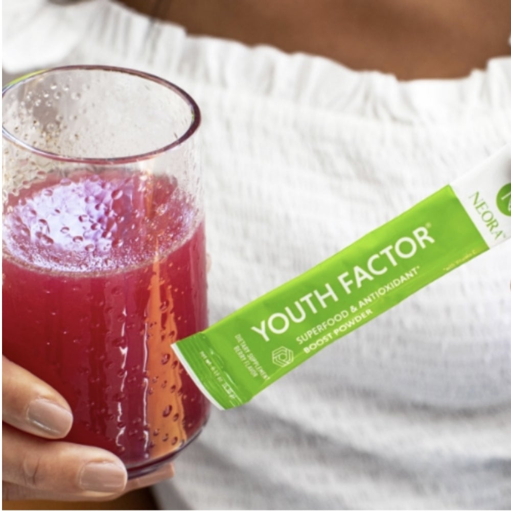 A woman holding a glass filled with red liquid & a packet of Neora Youth Factor.