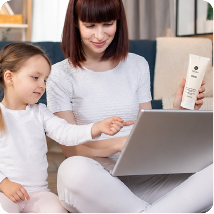 A mother holding a Firm Body Contour Cream tube while using her laptop with her daughter.