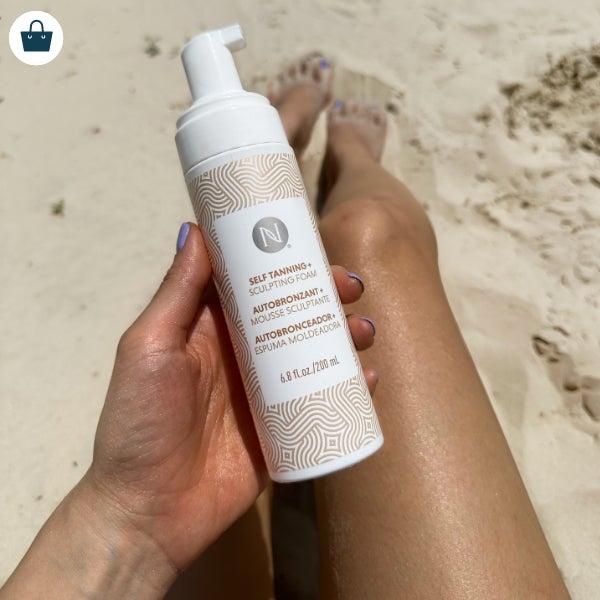 Woman holding 3-in-1 Self Tanning + Sculpting Foam while sitting on the beach.