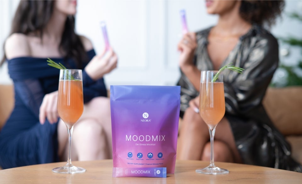 Two women holding MOODMIX packets in the background. In the foreground are two glasses mixed with MOODMIX and a MOODMIX bag in the middle.