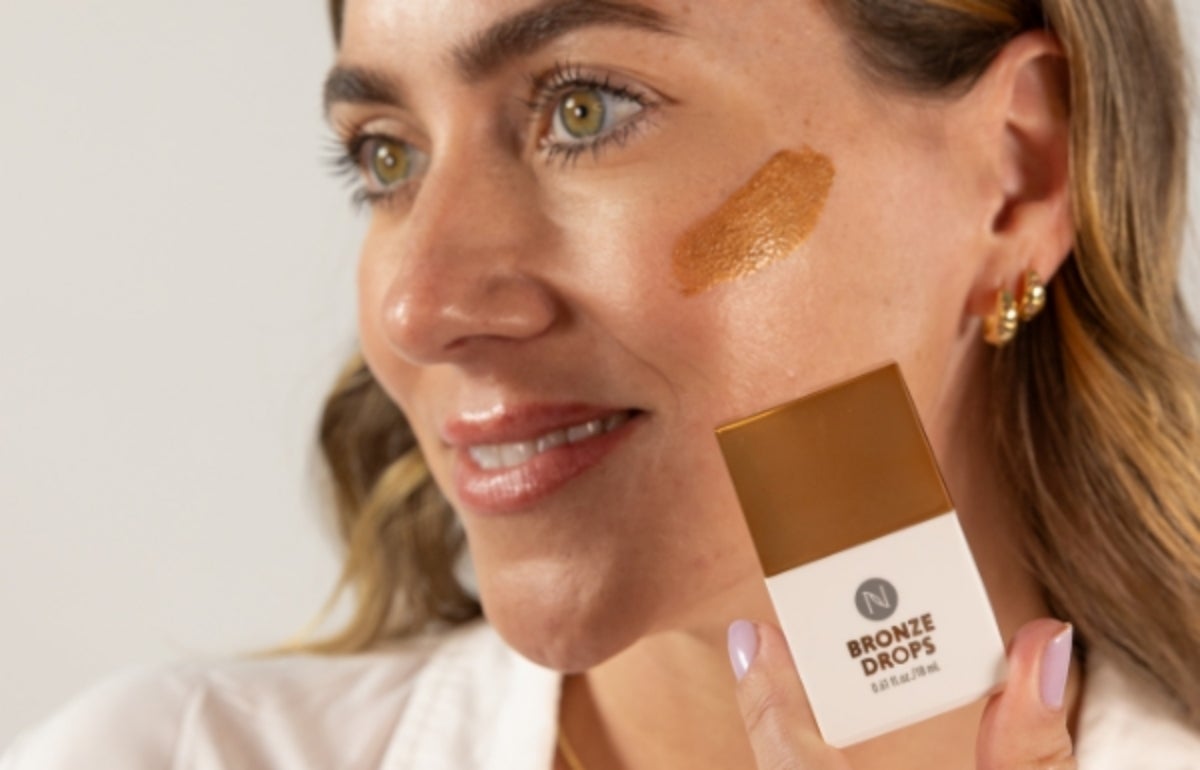 A woman holding a Bronze Drops container to her face with product smeared on her upper cheek.