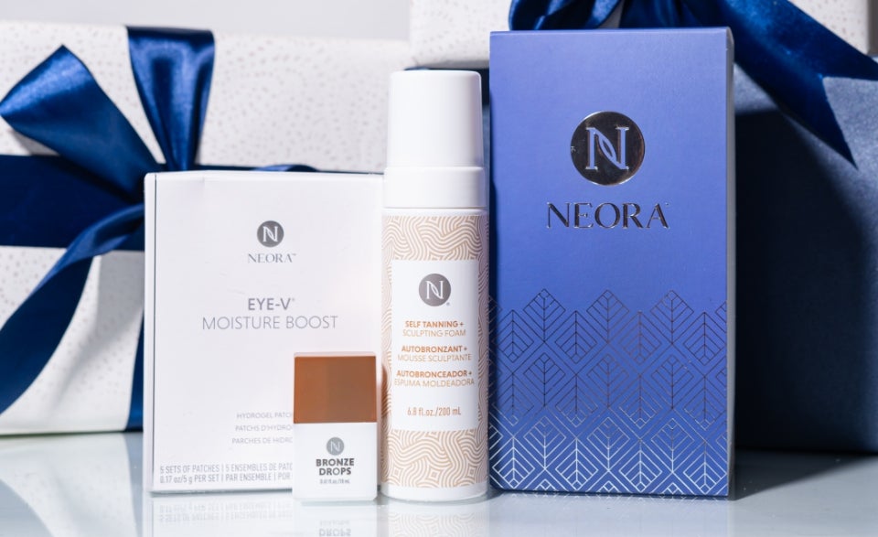 A bundle of Neora skincare products, which include Hydrogel Eye Patches, Bronze Drops, 3-in-1 Self Tanning + Sculpting Foam, and a Neora-branded gift box, surrounded by festive gift boxes.