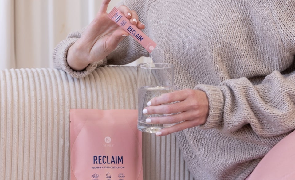A seated woman on a couch pouring a RECLAIM product sachet mixture into a drinking glass.