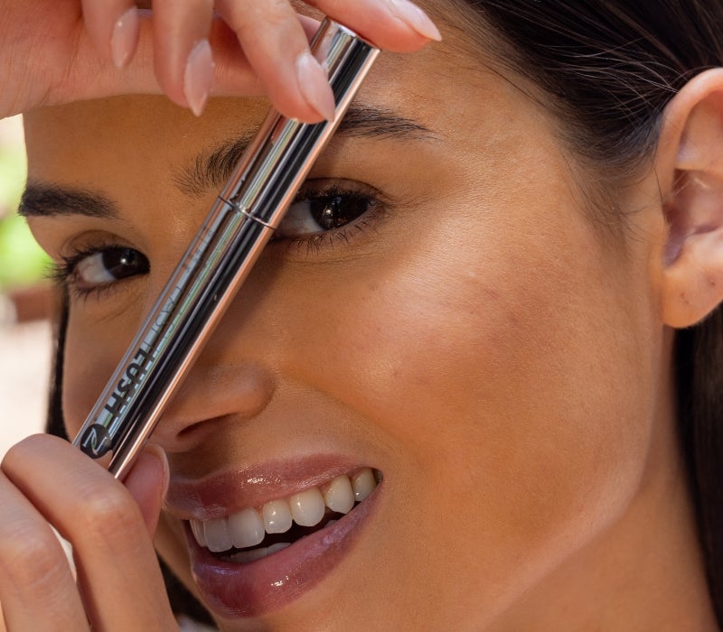 Alt text: A smiling woman holds up Neora’s LashLush 3-in-1 Lash & Brow Serum product over her face.