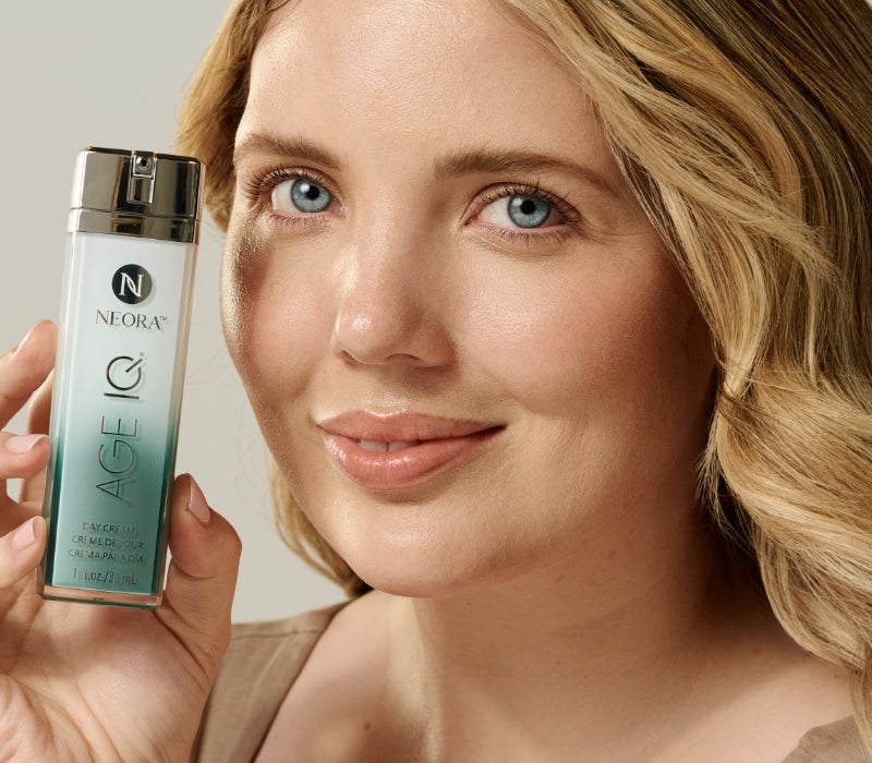 A woman holds up a bottle of Neora’s Age IQ Day Cream next to her face.