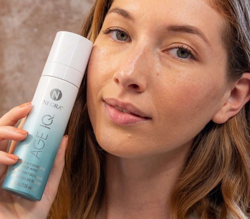 A woman holds up a bottle of Neora’s Age IQ® Double-Cleansing Face Wash next to her face.