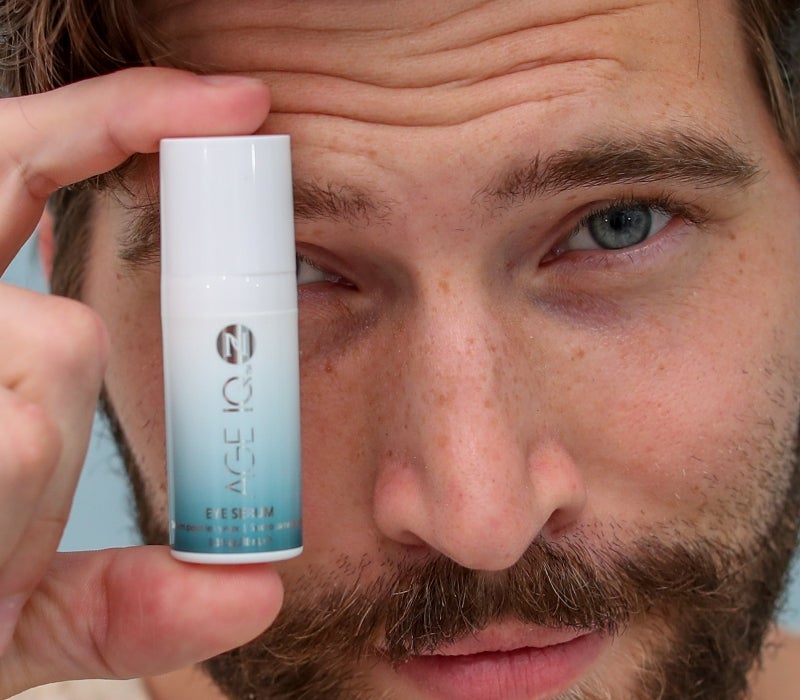 A man holds up a bottle of Neora’s Age IQ® Eye Serum next to his face.