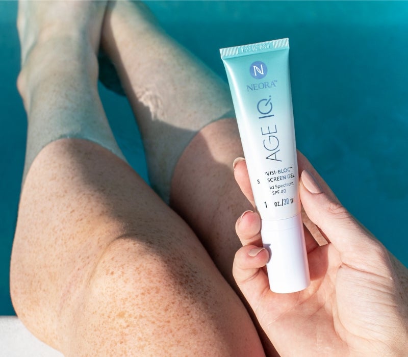 A person sits in a pool while holding up a tube of Neora’s Invisi-Bloc SPF40.
