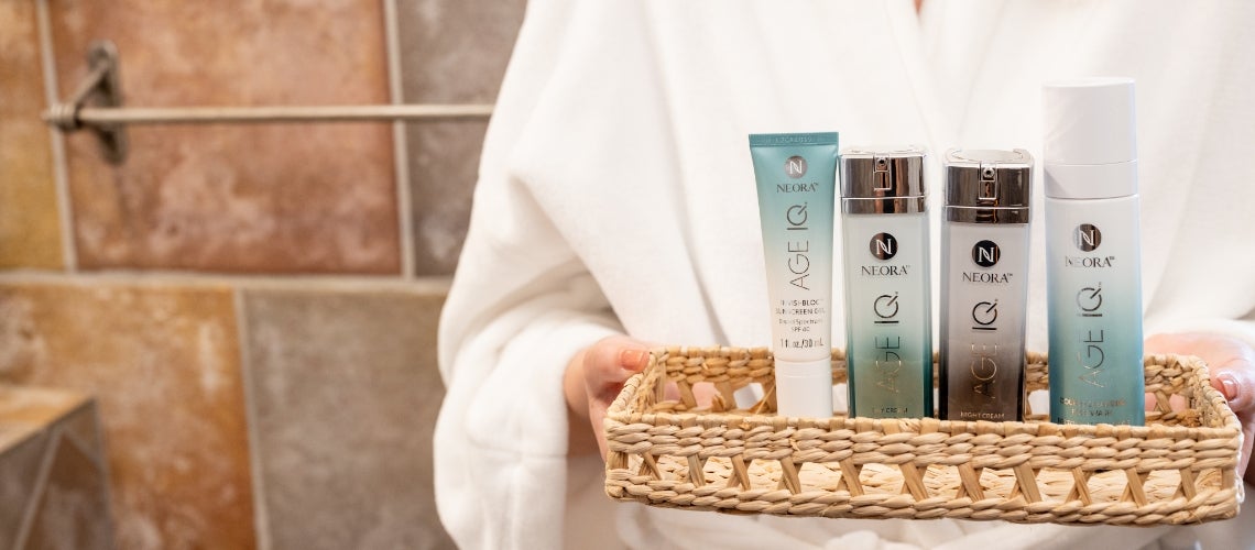 Spa scene with a person in a white robe holding a woven basket containing Neora skincare tubes and bottles.