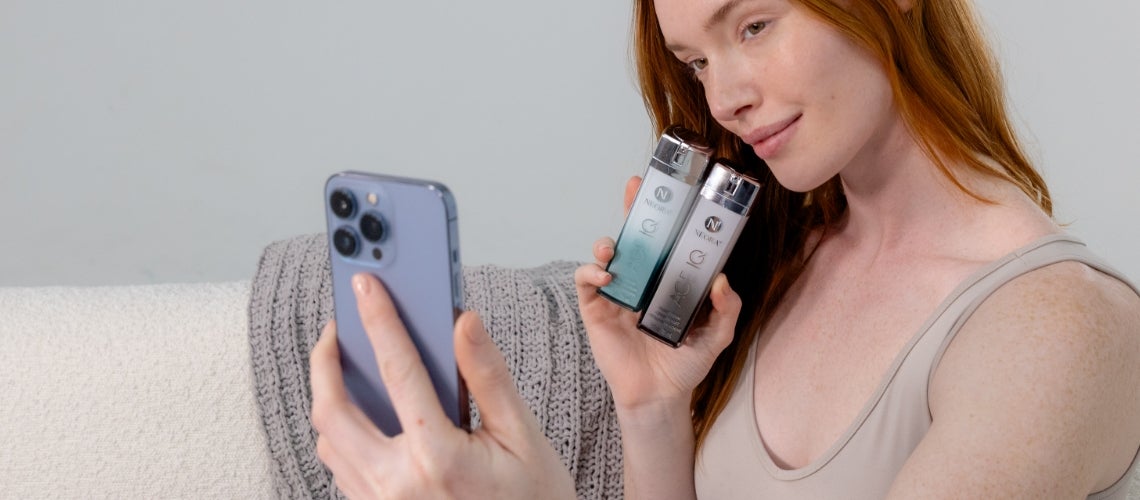 Red-haired woman on a sofa taking a selfie while holding two skincare bottles near her face and looking at her phone camera.