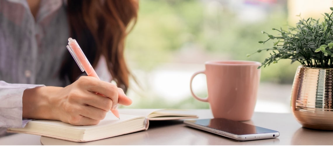 Person writes in a notebook with a pink pen at a desk, with a pink mug and a smartphone nearby.