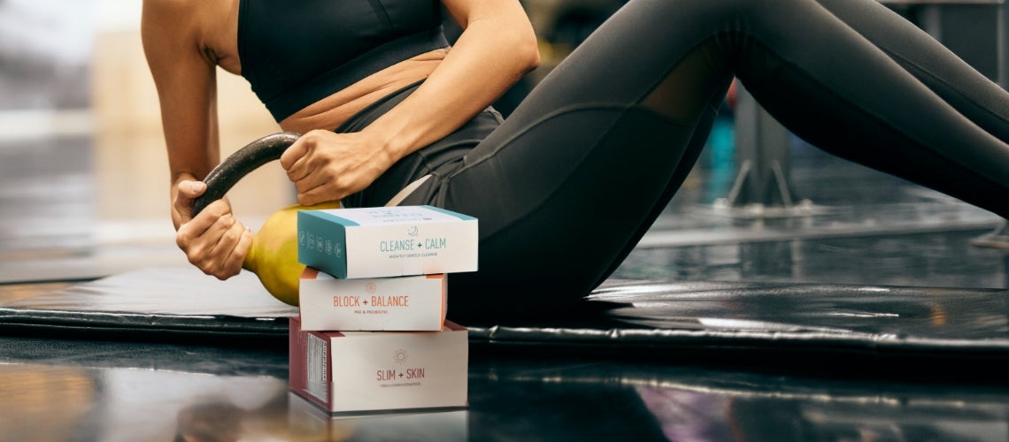 Fitness scene: person in black athletic wear doing a core crunch on a mat beside a kettlebell and stacked supplement boxes labeled Cleanse + Calm, Block + Balance, Slim + Skin.