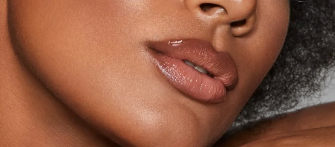 Close-up of a person's lips and chin with glossy, light brown lipstick and smooth skin tone.