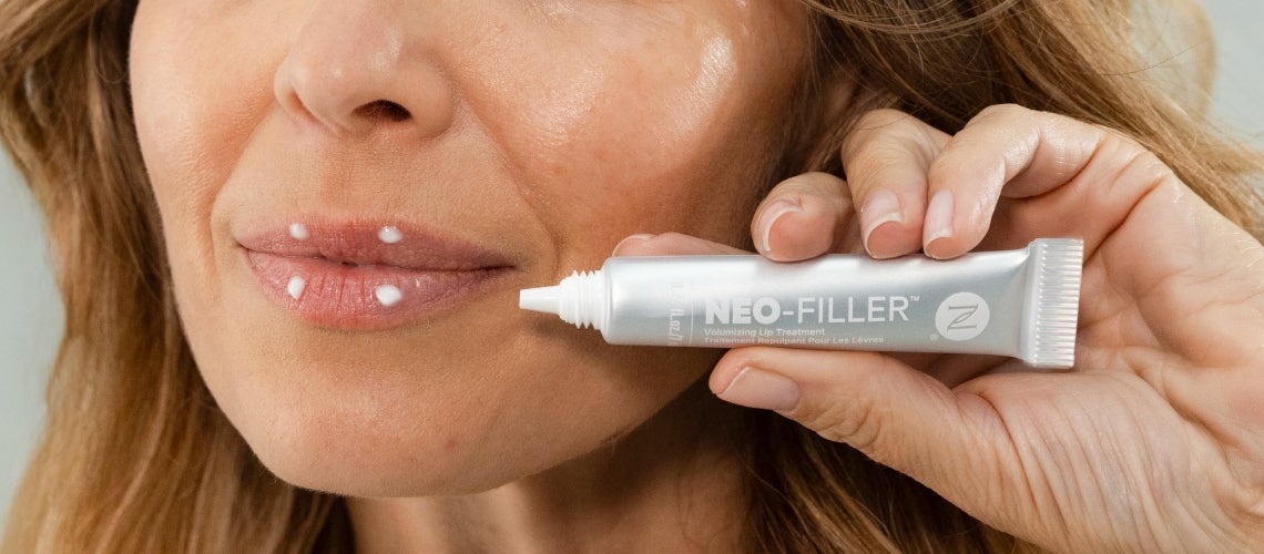 Close-up of a woman's lips with white cream dots, holding a tube labeled NEO-FILLER Volumizing Lip Treatment near her mouth.