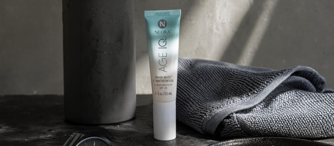 Tube of Neora Age sunscreen gel standing upright on a dark surface with a gray towel in the background.