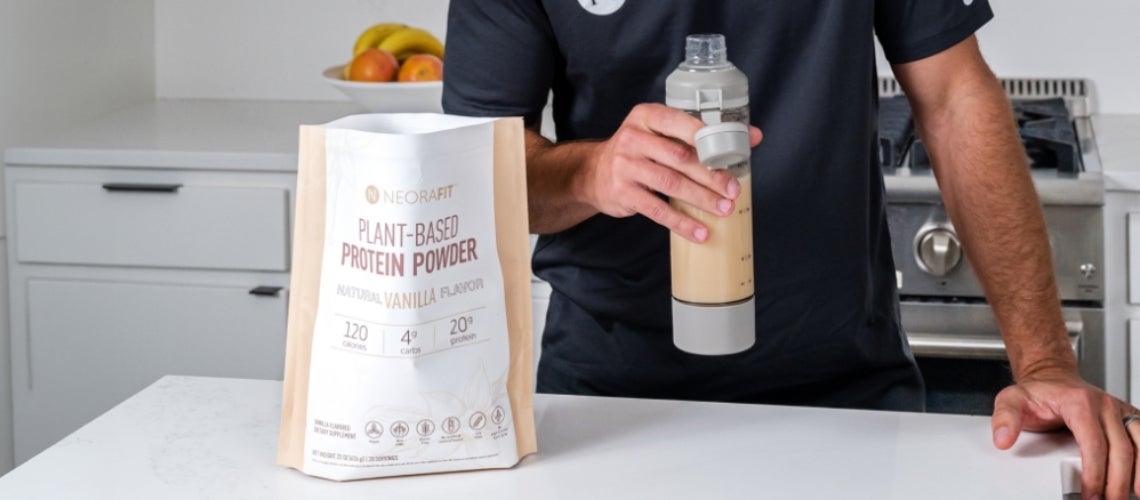 Person in a kitchen holds a shaker bottle beside a bag labeled Plant‑Based Protein Powder on a white counter.