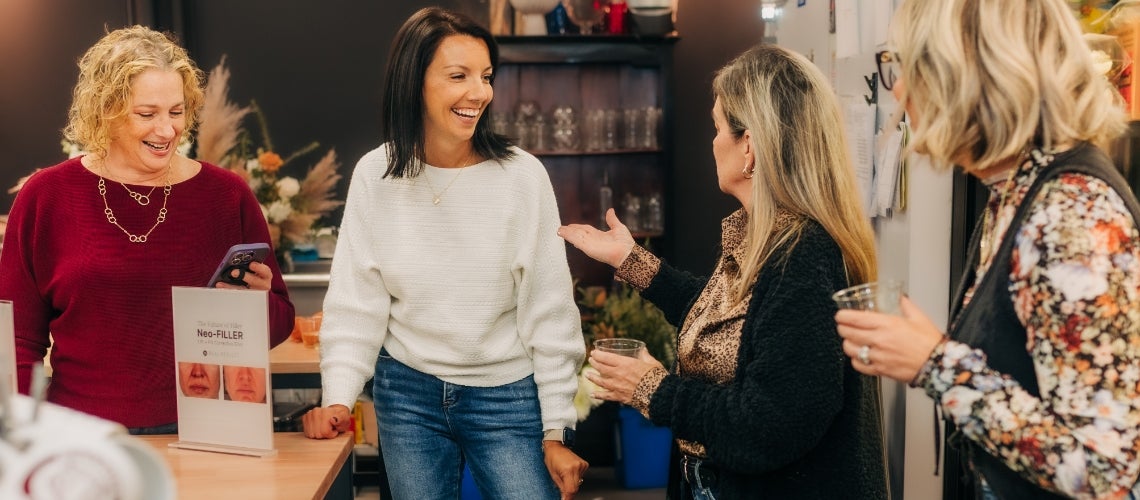 Four women chatting and smiling in a shop; one leans on the counter beside a Neo-Filler brochure on display.