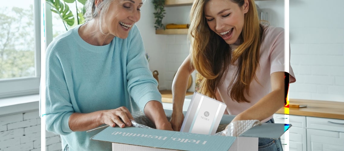 Two smiling women unpack a Neora skincare box in a bright, sunlit kitchen.