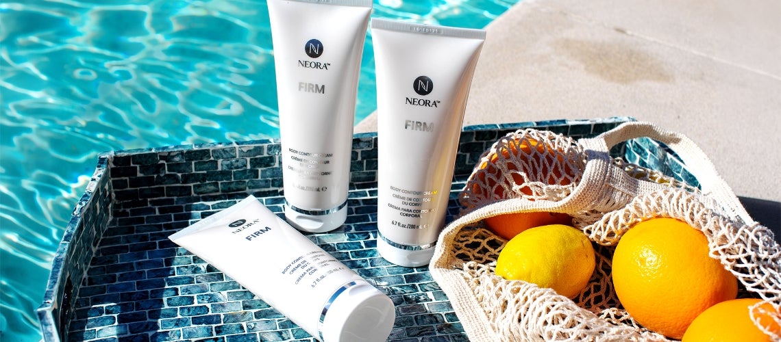 Neora Firm body contour cream tubes on a blue mosaic tray by a pool with a mesh bag of oranges nearby.