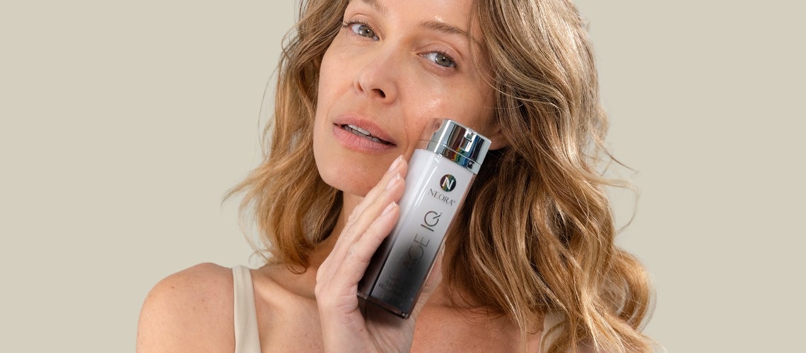 Woman applying a skincare serum from a white and silver bottle to her cheek against a beige background.