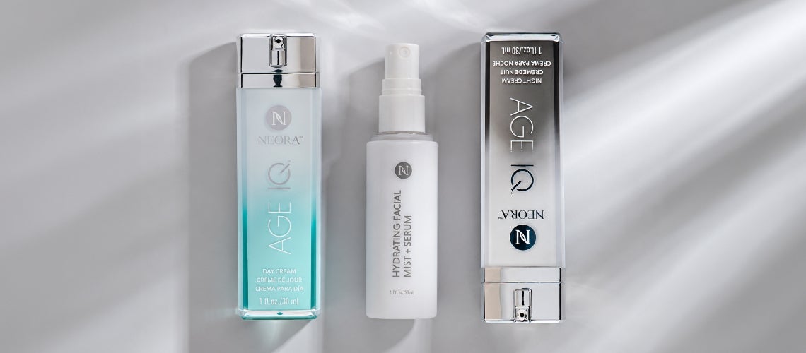 Three Neora Age IQ skincare products arranged on a light gray surface: a blue gradient day cream bottle, a white hydrating facial mist spray, and a boxed Age IQ product.