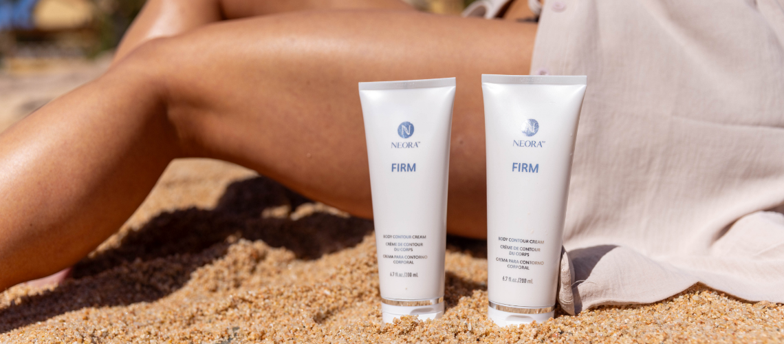 Two white tubes of Neora Firm body contour cream stand upright in sand at the beach, with a person’s leg in the background.