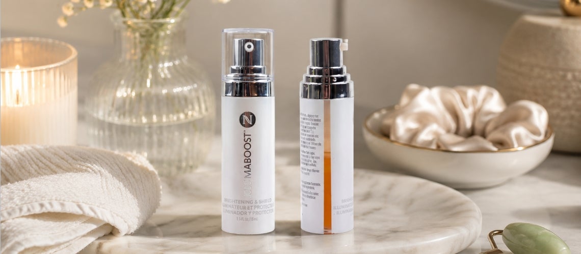 Two white skincare spray bottles with silver caps sit on a marble vanity, surrounded by spa props like a candle, vase, towel, and silk cloth.