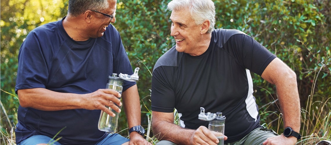 Two middle-aged friends in athletic wear sit on the grass, smiling and sharing a drink outdoors, with greenery in the background.