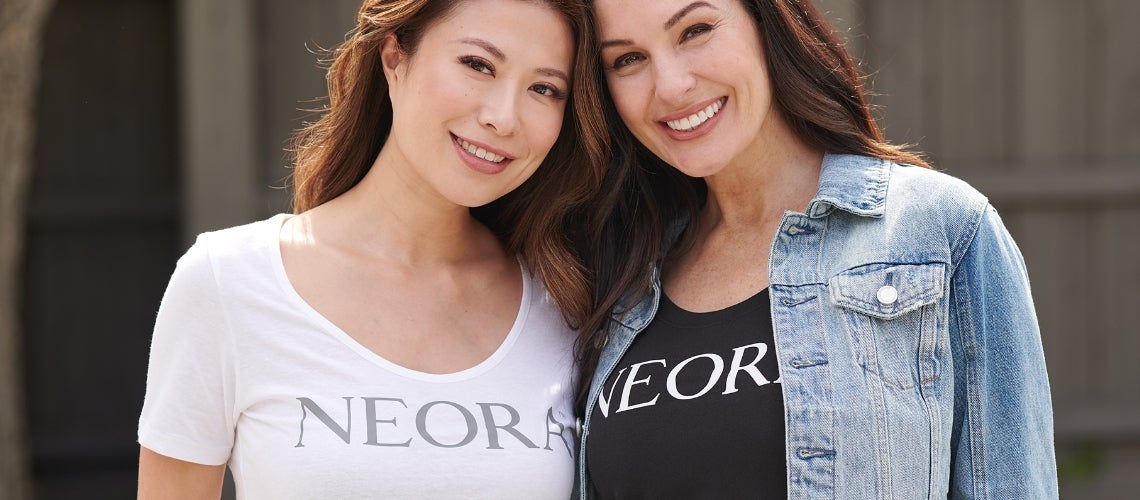 Two smiling women posing together for a photo outdoors, one in a white T-shirt and the other in a denim jacket.