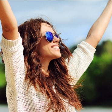 A woman wearing sun glasses and a white sweater embracing sunlight outside.