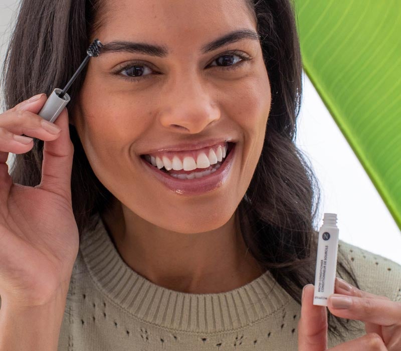 Woman holding Neora’s Hot List product, Brow Conditioning Serum, in her hand and applying it to her eyebrows while smiling