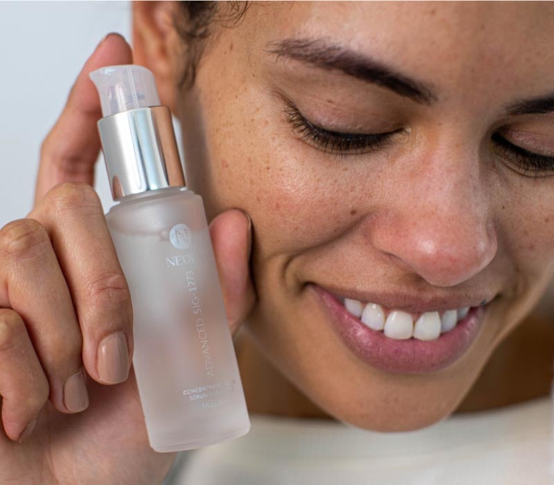 Woman holding Neora’s Advanced SIG-1273 Concentrated Serum 
                        in her hand next to her face while smiling 